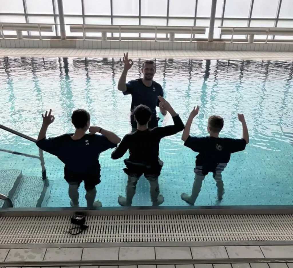 Tauchlehrer und drei Kinder stehen im Schwimmbecken und zeigen stolz das OK-Zeichen