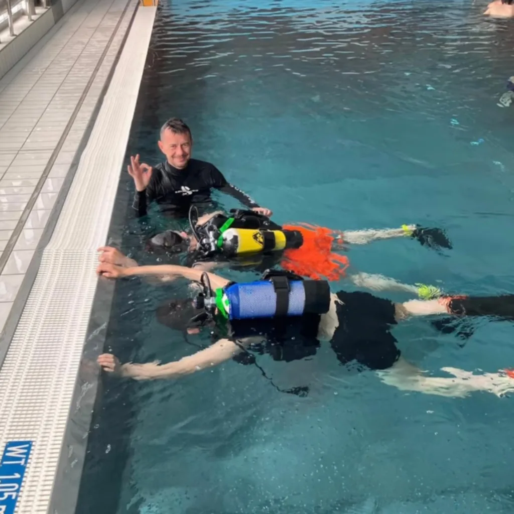 Tauchlehrer zeigt das OK-Zeichen, während zwei Kinder mit kleinen Tauchflaschen an der Wasseroberfläche schweben